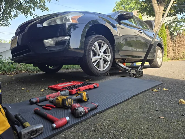 A car raised on jack stands in a driveway, tools laid out on rubber mats